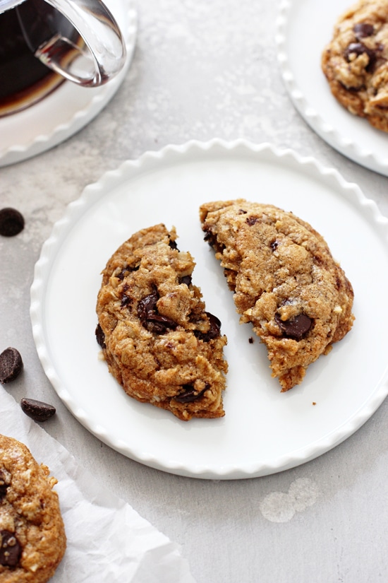 A Coconut Oil Oatmeal Cookie split in half on a white plate with coffee and more cookies in the background.