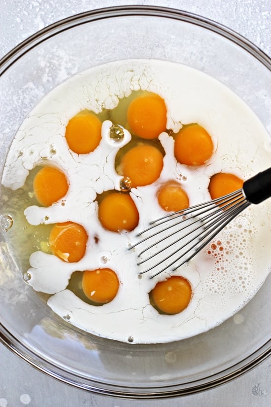 A glass mixing bowl with eggs, almond milk and a whisk.