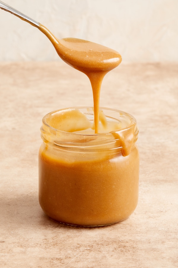 A jar of Dairy Free Caramel Sauce with some being drizzled with a spoon.